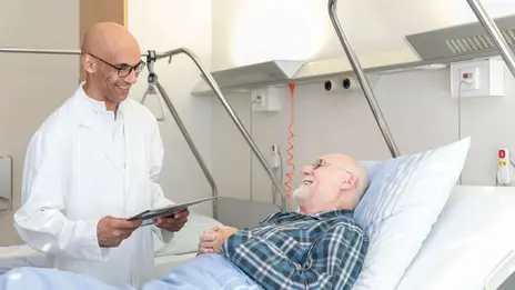 Ein Arzt spricht mit einem männlichen Patienten im Bett. Die Stimmung ist freudig. 
